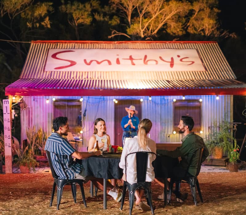 Four people sitting at a dinner table outside a rustic corrugated iron building with "Smithy's" sign at night.