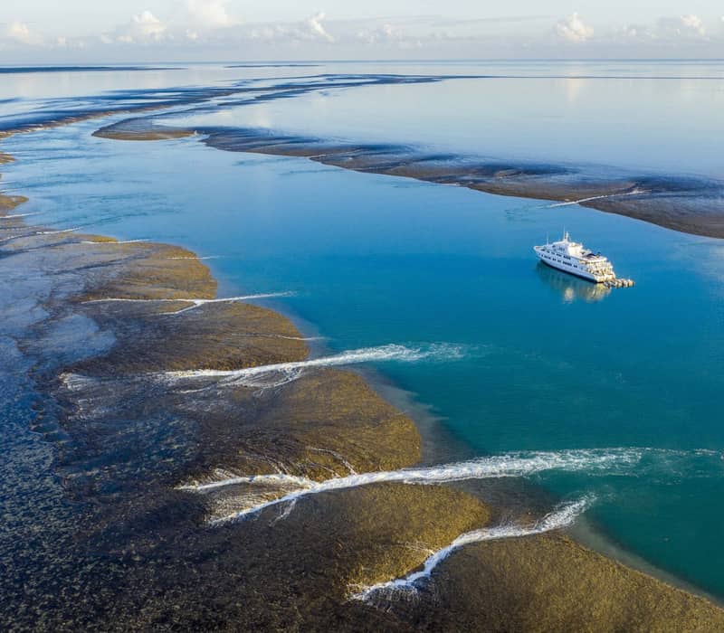 An aerial shot showing a white luxury cruise ship in a deep blue channel between vast, exposed dark coral reefs with water cascading off the edges as the tide recedes.