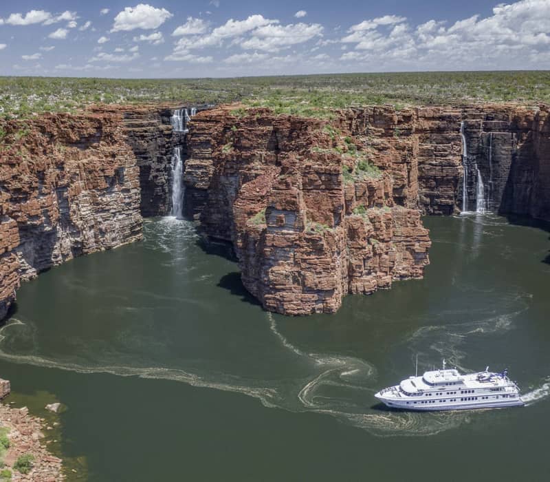 A high-angle aerial view of a white cruise ship navigating near several large waterfalls cascading down high, red sandstone cliffs into a green river.