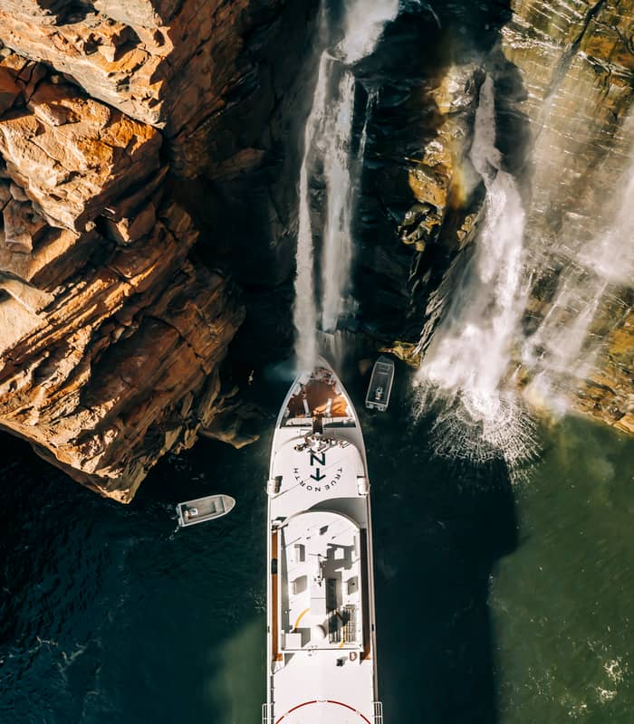 A vertical top-down aerial shot shows a white luxury cruise ship True North with its bow positioned directly under a large waterfall cascading down a high, jagged red rock cliff. Two small gray adventure boats are seen in the dark water nearby.