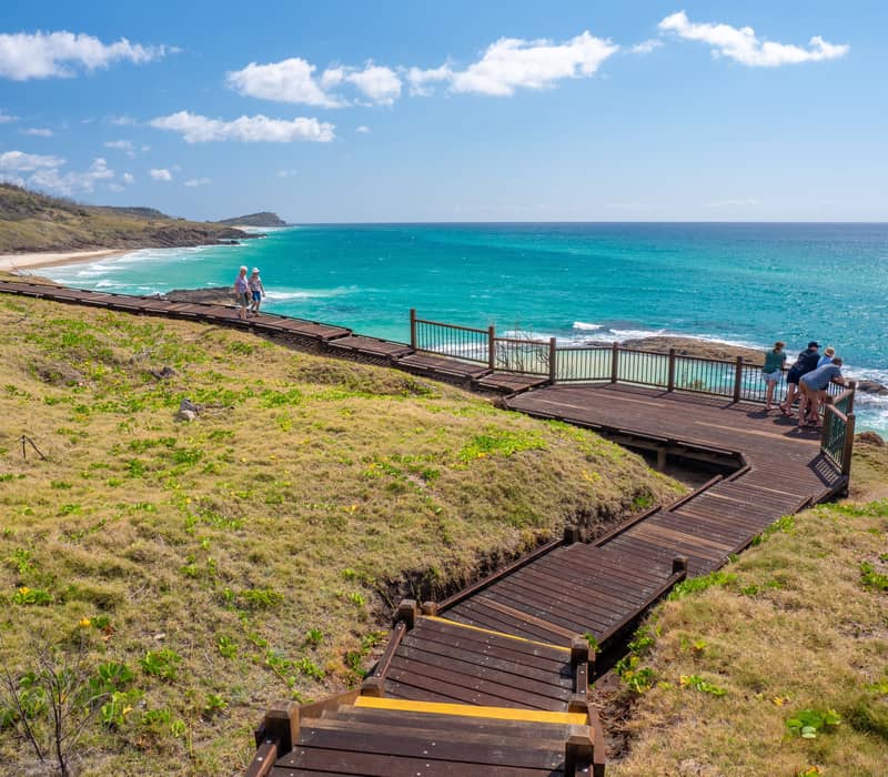 A traveler walks along the wooden boardwalk overlooking the Champagne Pools, K'gari's only safe saltwater swimming spot where waves crash over volcanic rocks to create a natural "bubbly" spa.