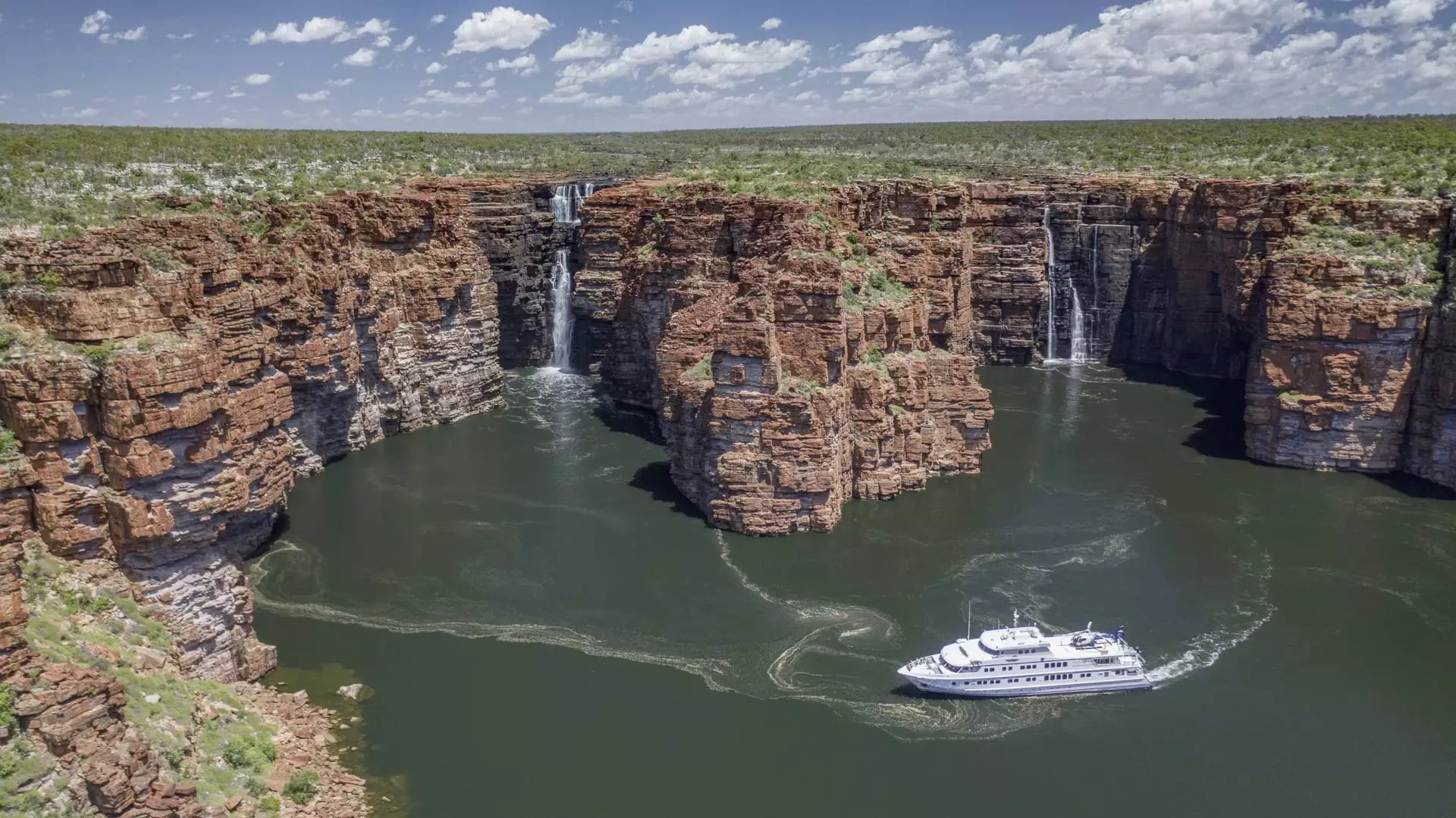 A high-angle aerial view of a white cruise ship navigating near several large waterfalls cascading down high, red sandstone cliffs into a green river.