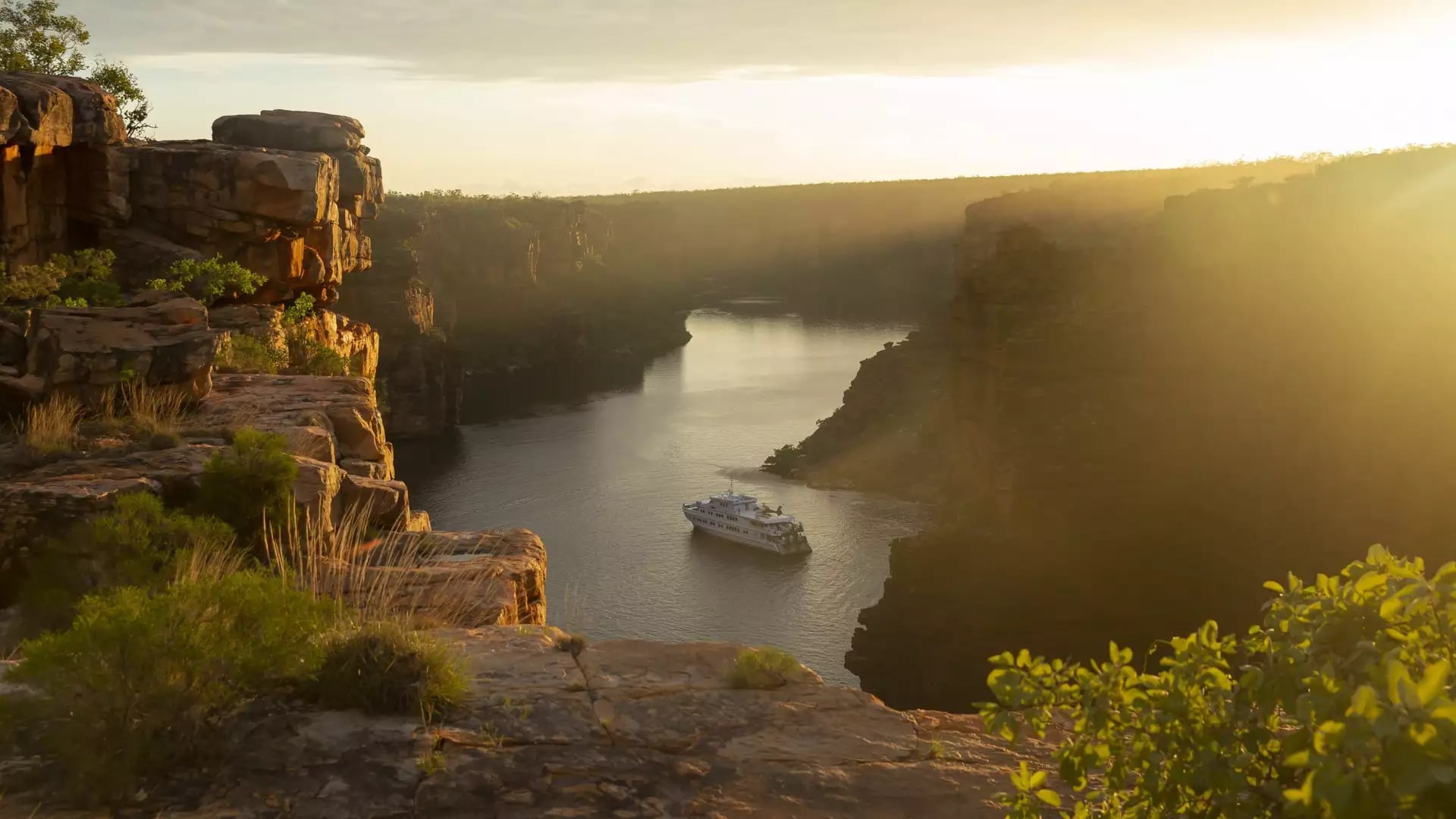 A wide aerial shot of a white luxury cruise ship anchored in a narrow, calm river gorge at sunset, with high, jagged orange rock walls and golden light reflecting off the water.