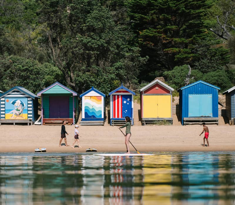 A woman on a stand-up paddleboard in calm water with a row of brightly painted beach huts on the shore.