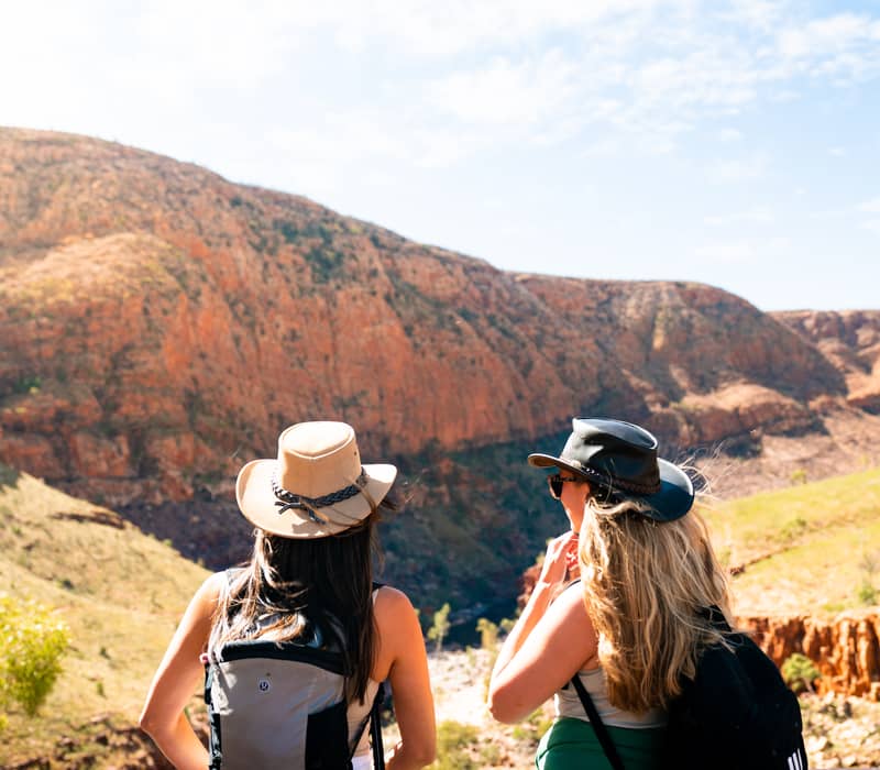 Rear view of two women wearing sun hats and backpacks looking out over a vast valley with towering orange rock walls under a bright blue sky.