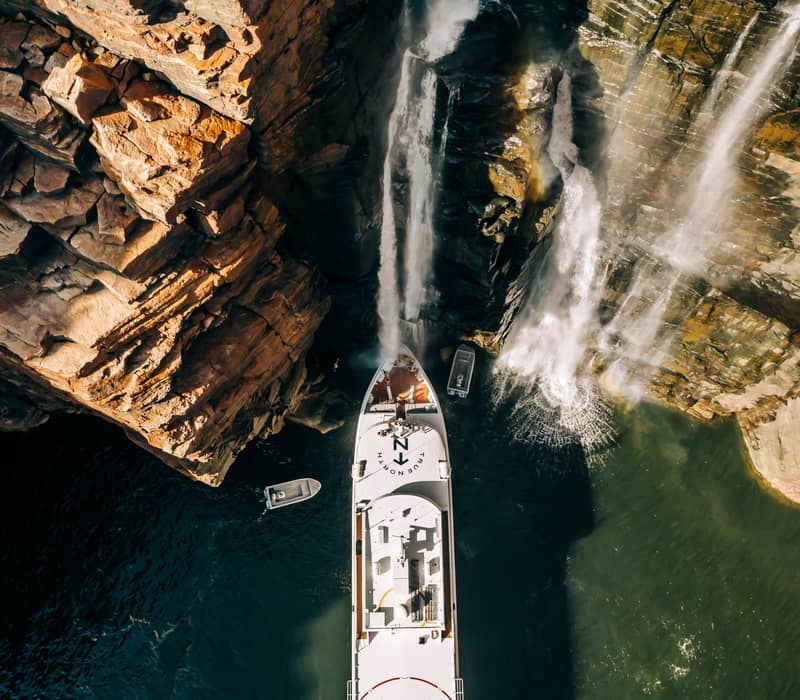 A vertical top-down aerial shot shows a white luxury cruise ship True North with its bow positioned directly under a large waterfall cascading down a high, jagged red rock cliff. Two small gray adventure boats are seen in the dark water nearby.