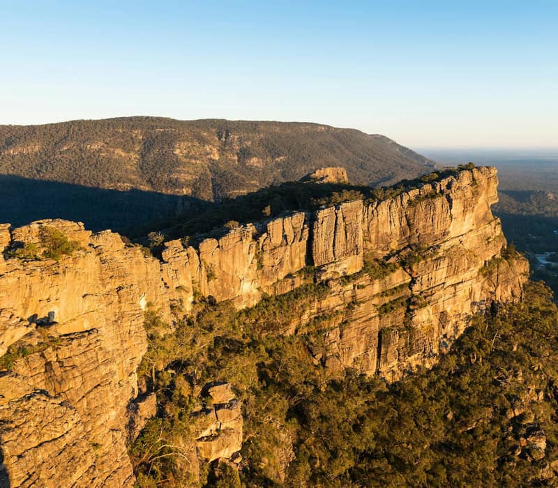 Golden sunlight on rocky cliffs and forested mountains of the Grampians at sunset.