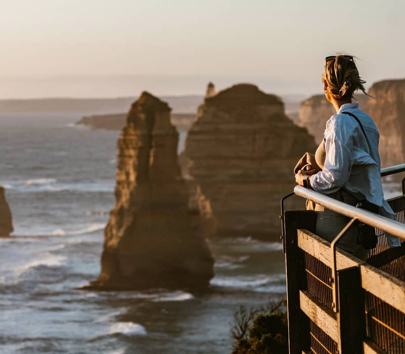 Woman standing at a coastal lookout railing overlooking the Twelve Apostles rock stacks during sunset on the Great Ocean Road.