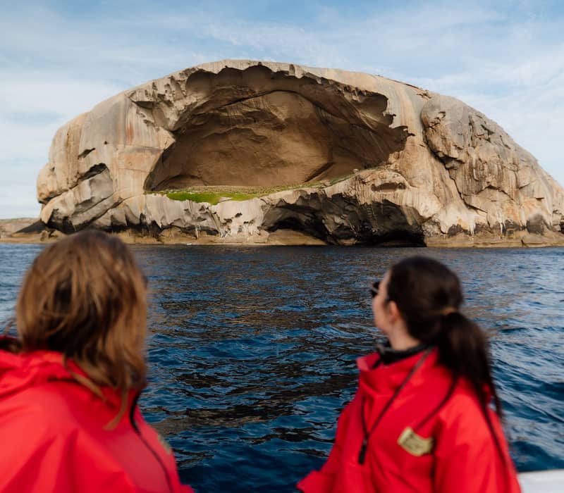 Two people in red jackets on a boat looking at a giant, skull-shaped rock island in the sea.