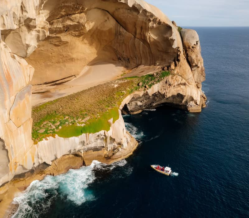 Aerial view of a small yellow boat next to a massive, hollowed-out granite island in the deep blue ocean.