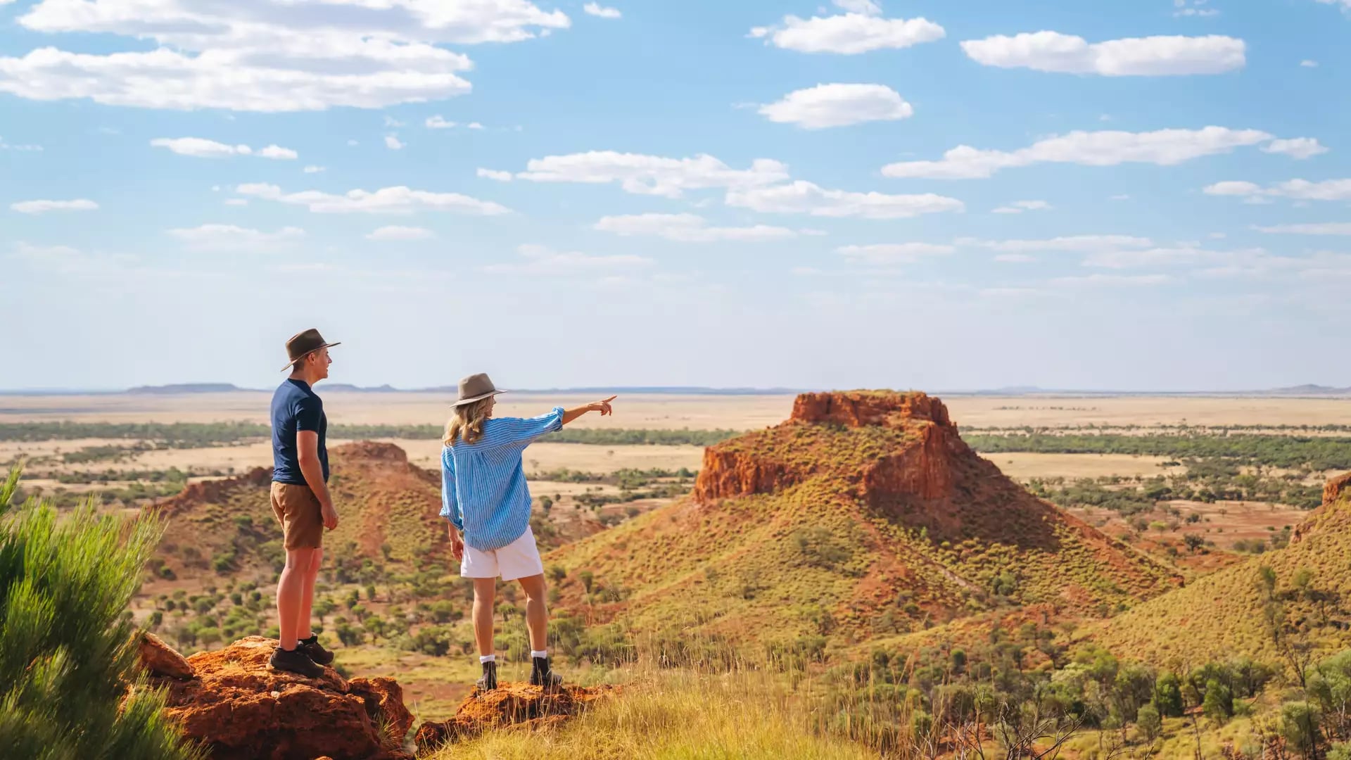 Two people standing on a rocky outcrop looking out over a vast outback landscape with red mesas.