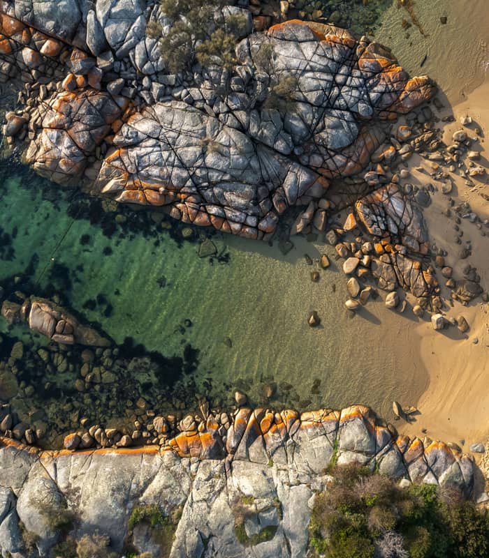 direct top-down aerial photograph of a secluded cove in the Bay of Fires, featuring crystal-clear turquoise water, white sand beach with footprints, and large granite boulders splashed with vibrant orange lichen.