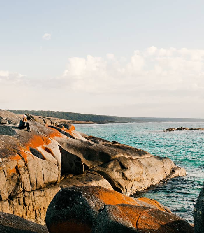 direct top-down aerial photograph of a secluded cove in the Bay of Fires, featuring crystal-clear turquoise water, white sand beach with footprints, and large granite boulders splashed with vibrant orange lichen.
