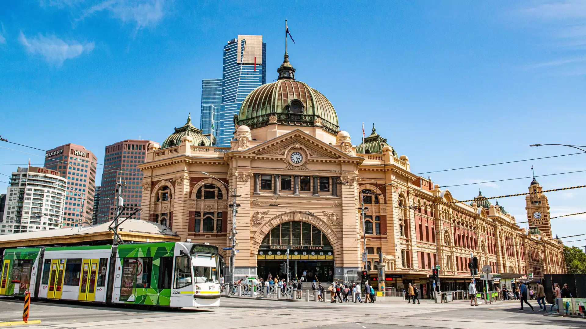 A green and white Melbourne tram driving past the historic Flinders Street Station under a blue sky.