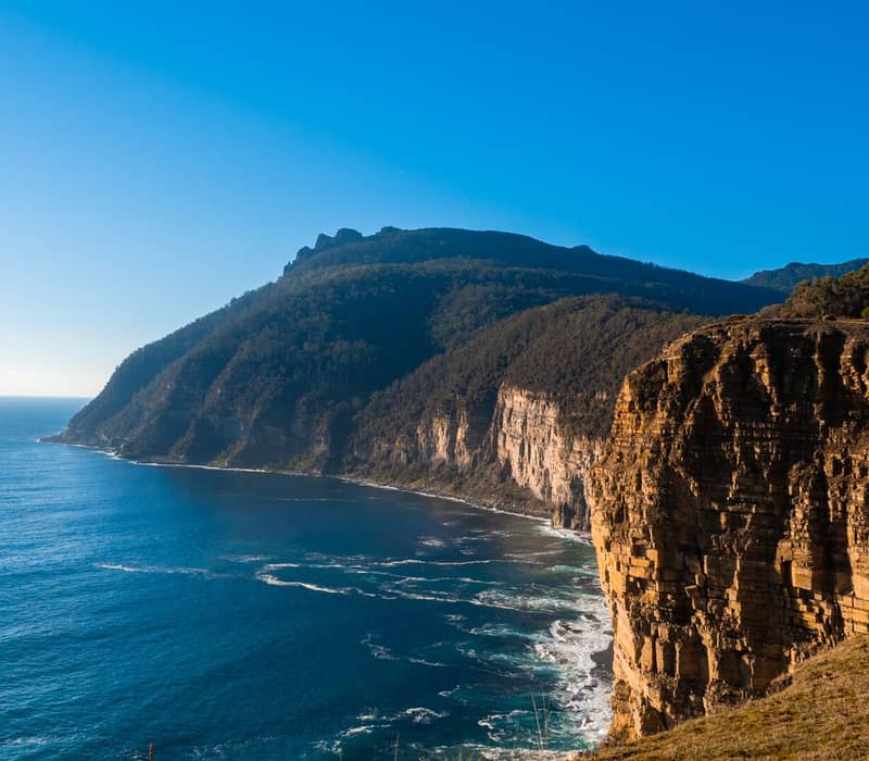 A scenic coastal view featuring high, steep yellow and brown rock cliffs dropping into a vibrant blue sea under a clear blue sky.