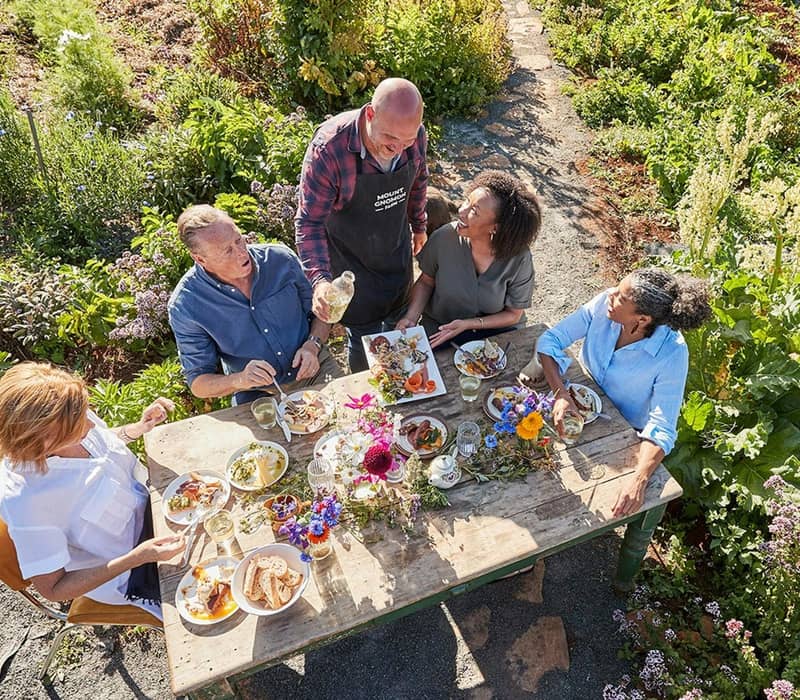 A group of people sharing a meal and wine at a rustic wooden table in a blooming kitchen garden under the sun.