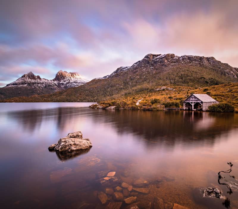 Long exposure of Dove Lake boat shed at sunset with pink clouds and Cradle Mountain reflections in Tasmania.