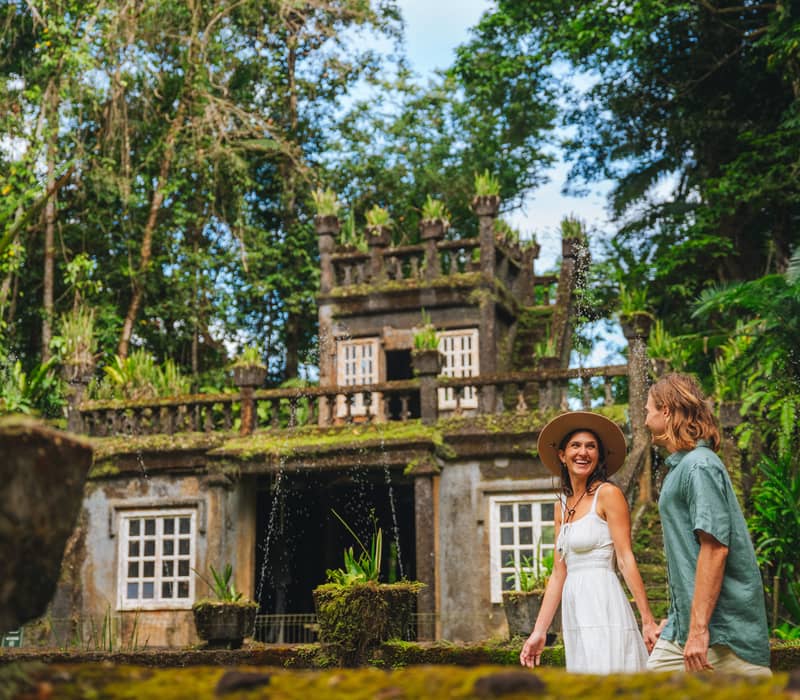 A couple holding hands walks past the moss-covered castle ruins and tropical gardens of Paronella Park in Mena Creek.
