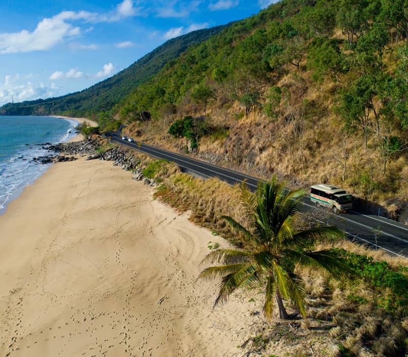 Aerial view of a coastal road winding between a lush green mountain and a sandy beach with turquoise water.