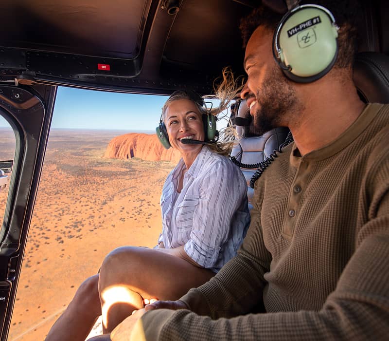 Interior of a helicopter showing a smiling couple looking out at Uluru in the distance.