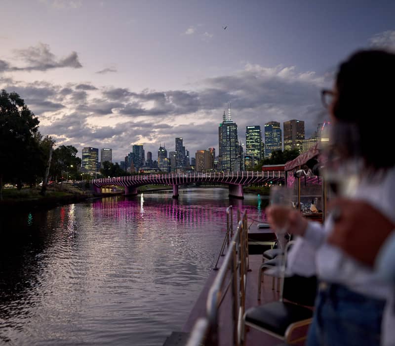 View from the deck of a boat on the Yarra River at dusk looking toward the illuminated Melbourne CBD.