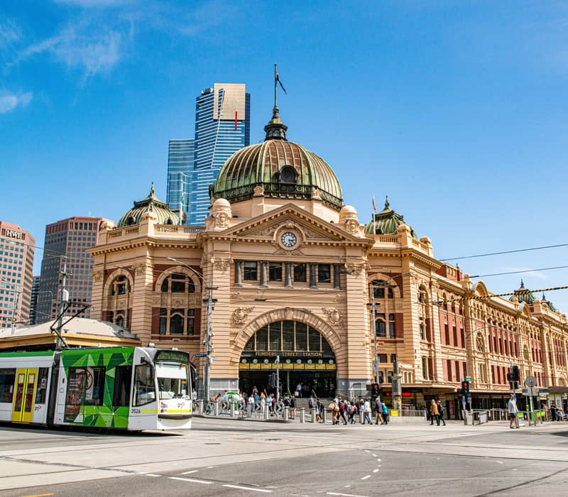 A green and white Melbourne tram driving past the historic Flinders Street Station under a blue sky.