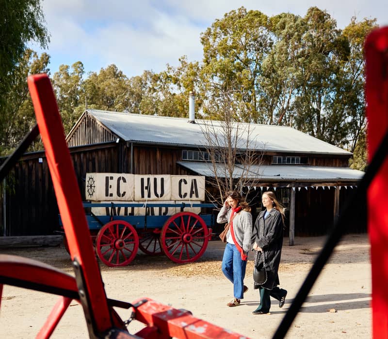 Two women walking past a vintage horse-drawn wool wagon at the Port of Echuca heritage site.