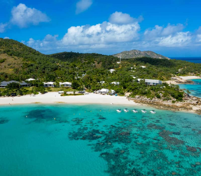 Aerial view of Lizard Island Resort beach with clear turquoise water, white sand, boats, and green hills in the background.