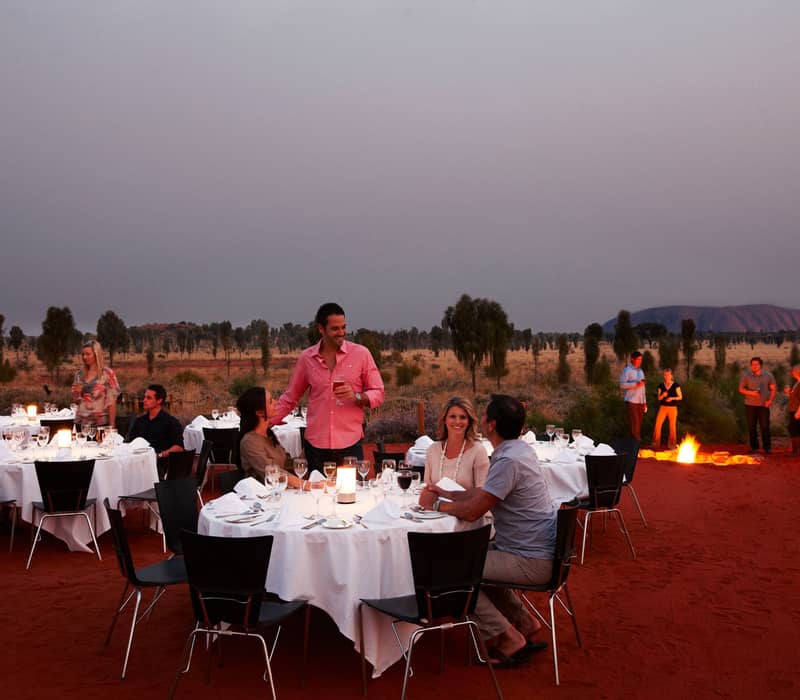 Couples sitting at round tables on red sand at dusk, being served drinks by a waiter with Uluru in the background.