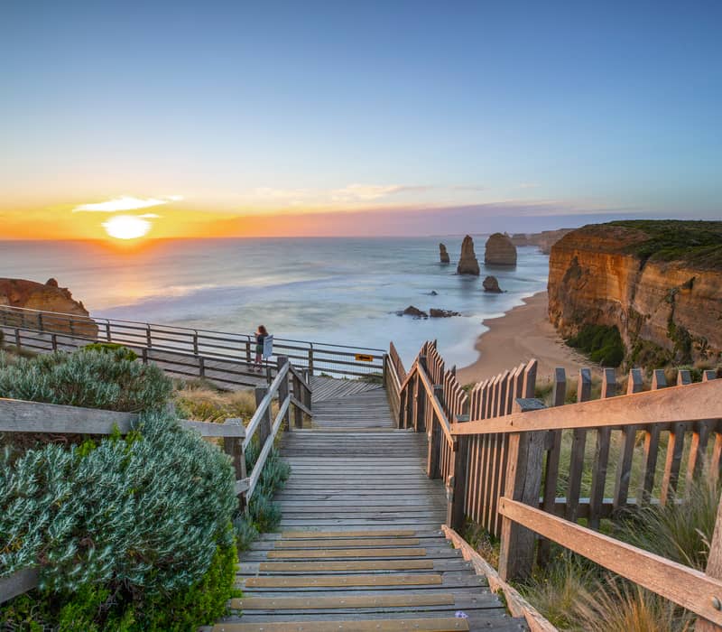 A wooden staircase and boardwalk overlooking the Twelve Apostles limestone stacks at sunset.