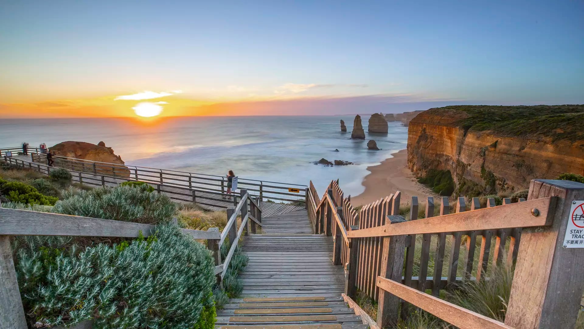 A wooden staircase and boardwalk overlooking the Twelve Apostles limestone stacks at sunset.