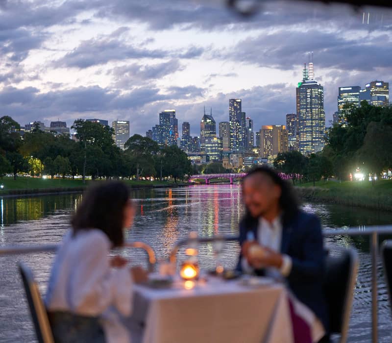 A couple dining at a white-clothed table on a boat with the Melbourne city lights reflecting in the river.