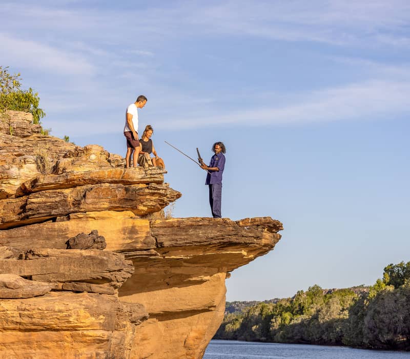 An Aboriginal guide stands on a dramatic sandstone ledge above the calm East Alligator River, holding a traditional spear and demonstrating its use to a man and woman standing nearby in Kakadu.