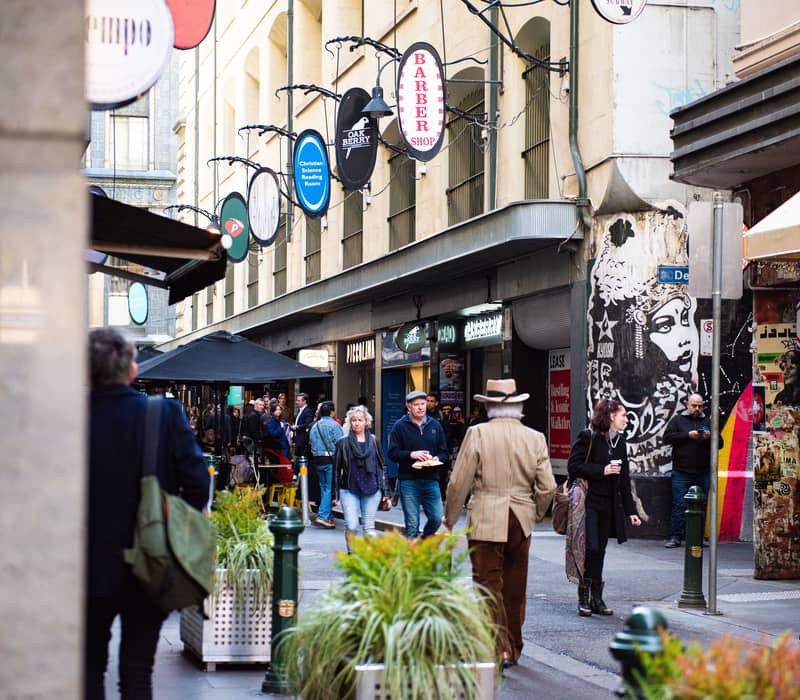 People walking through a busy Melbourne laneway filled with cafes, shops, and colorful street art.