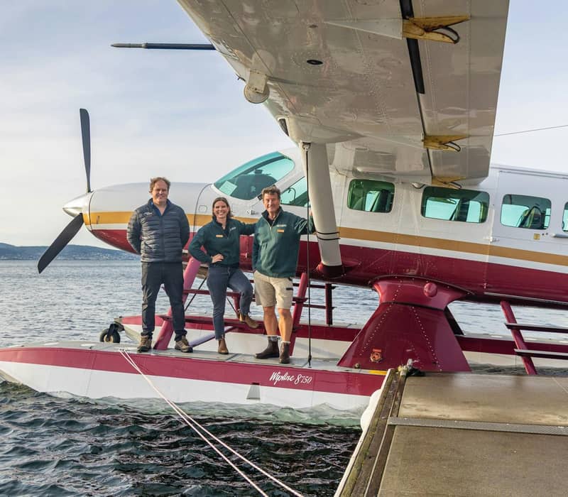 Three people pose on the floats of a docked seaplane over water with land visible in the background.
