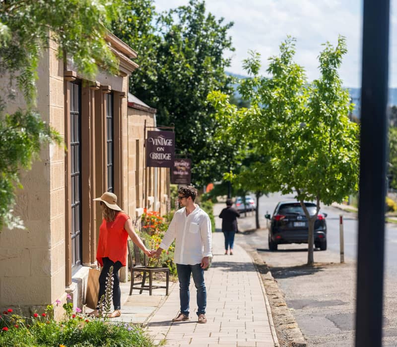 A man and woman holding hands while walking past vintage sandstone storefronts on a sunny day in Richmond, Tasmania.