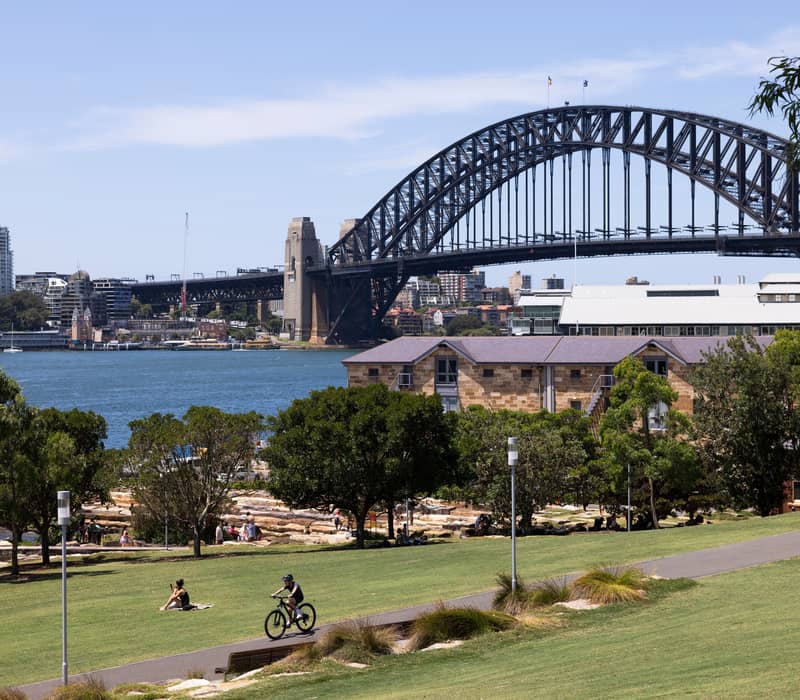 A cyclist on a path in Barangaroo Reserve park with the Sydney Harbour Bridge in the distance.