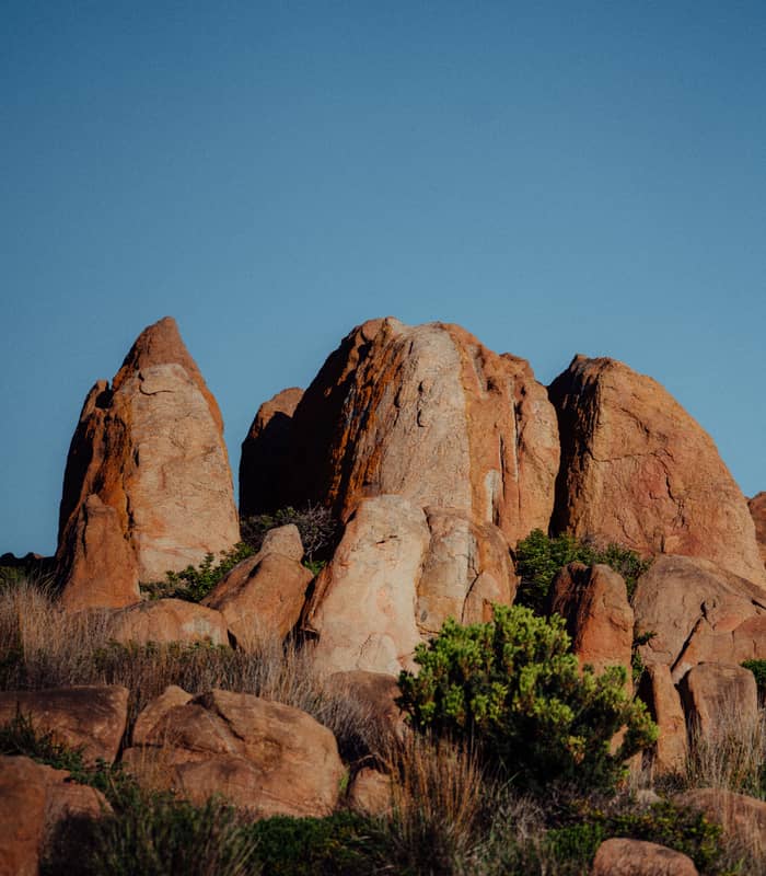 Large, tan and orange rock formations with some green vegetation at the base, under a clear blue sky.