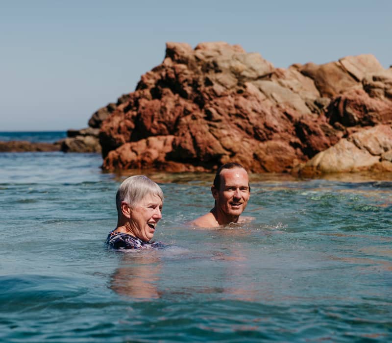 Two smiling older adults swimming happily in the ocean near large reddish-brown rocks of the Injidup Natural Spa