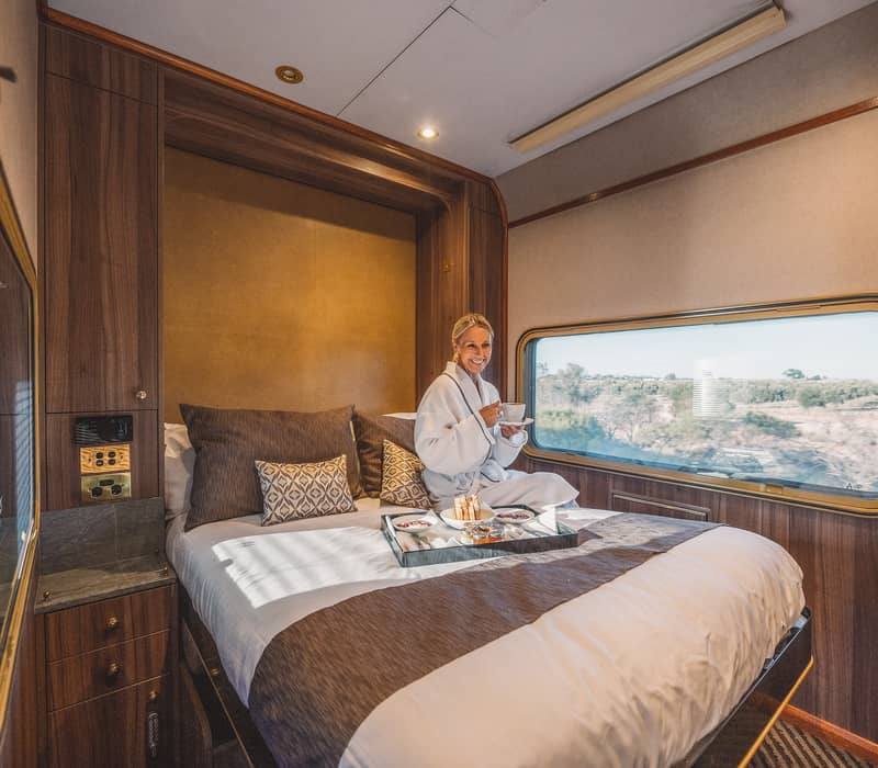 Woman in a white robe drinking tea on a large bed inside a luxury train cabin looking out the window.