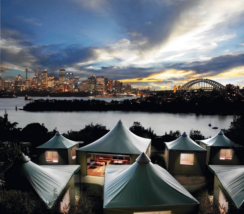 Night view of glamping tents at Taronga Zoo with the illuminated Sydney city skyline and Harbour Bridge in the background.
