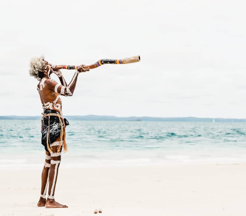 A profile shot of an Indigenous Australian man with white body paint and grey hair playing a long, decorated didgeridoo on a white sand beach.
