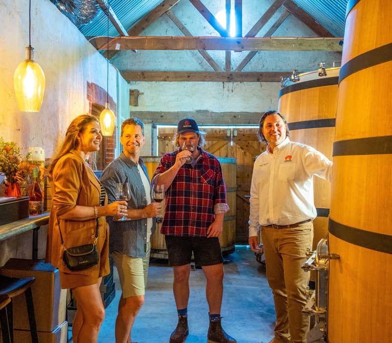 People enjoy a wine tasting inside a rustic Australian cellar with large wooden vats.