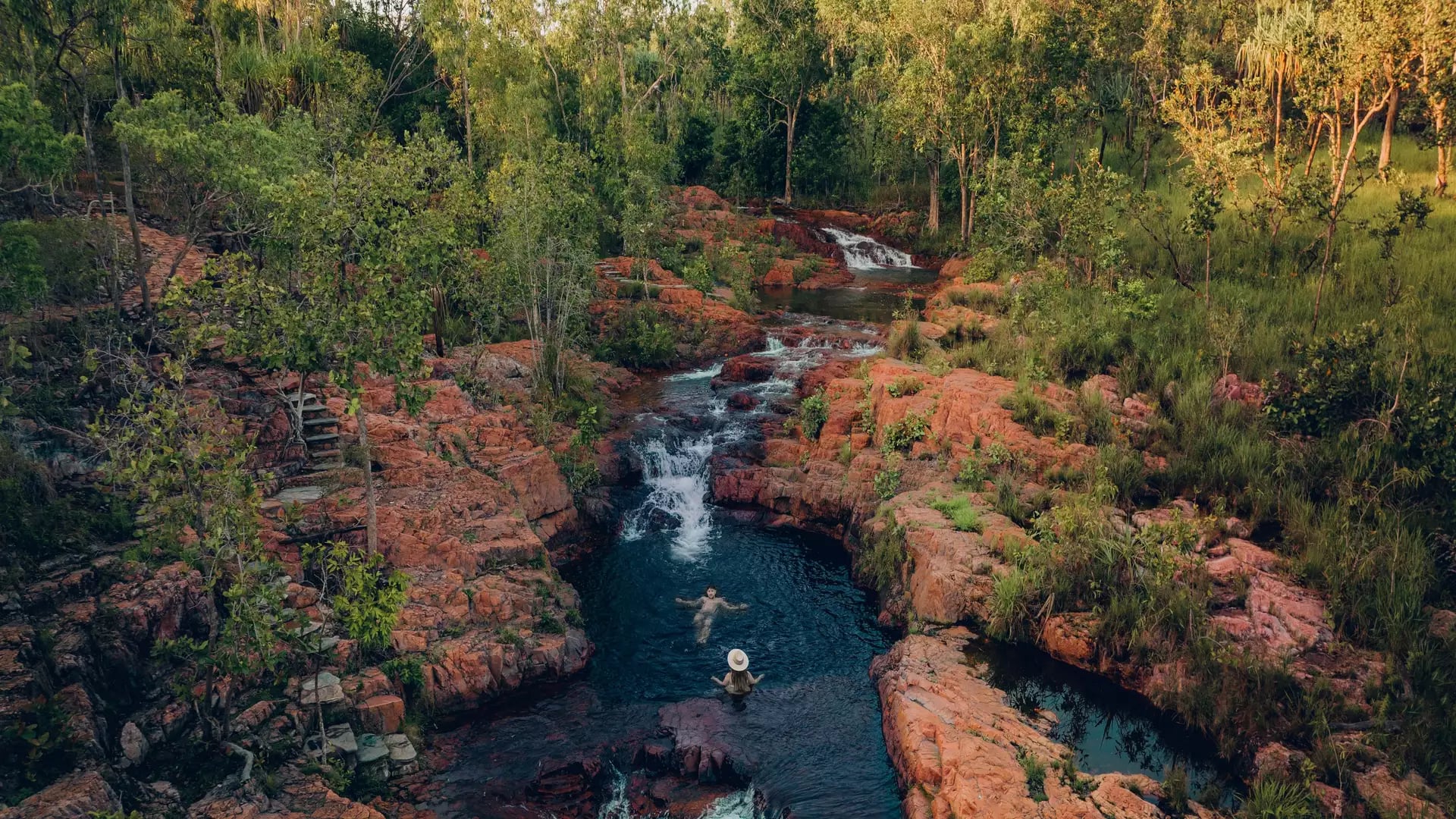 People swimming in tiered natural waterfalls and rock pools surrounded by green forest in Litchfield National Park.