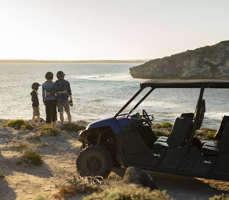 A family of three in helmets standing next to a blue off-road buggy looking out at the ocean waves.