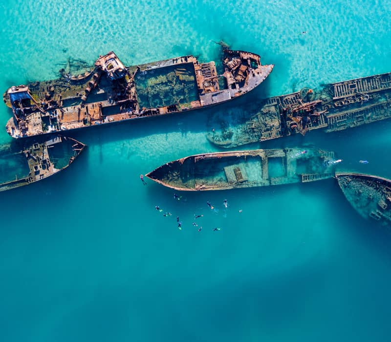 From a top-down aerial view, snorkellers swim through the clear blue water surrounding the rusted hulls of the Tangalooma Wrecks on Moreton Island, Queensland.