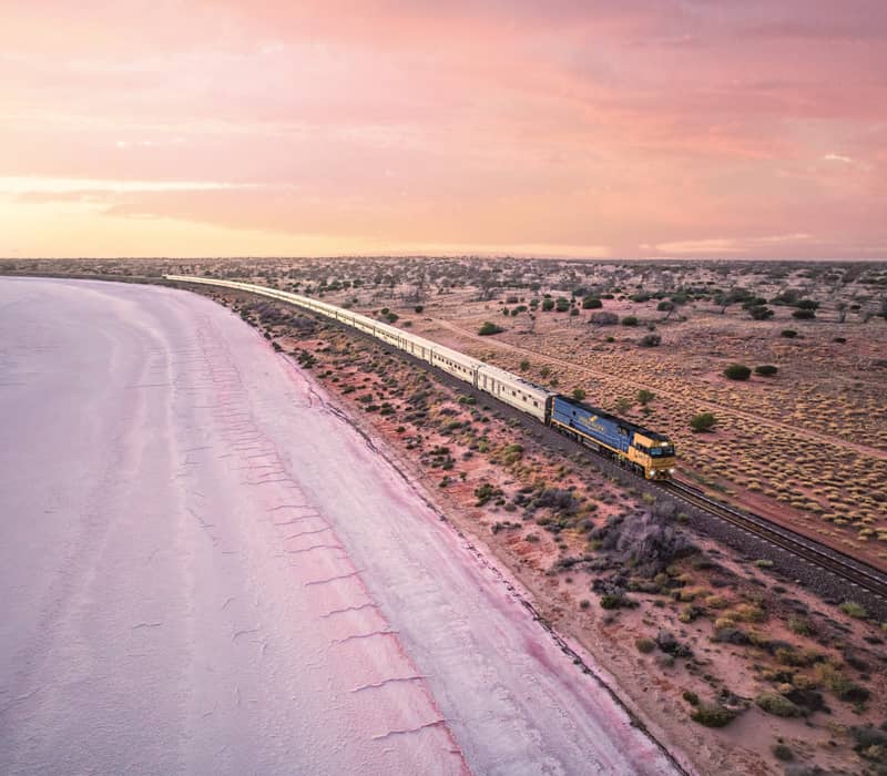 High-angle view of the long Indian Pacific passenger train traveling alongside a large white salt lake under a pastel sunset sky.