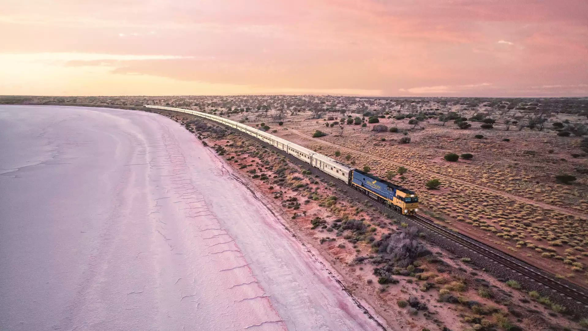 High-angle view of the long Indian Pacific passenger train traveling alongside a large white salt lake under a pastel sunset sky.