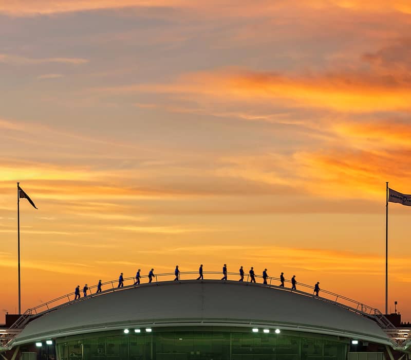 Silhouette of a group of people walking across the curved stadium roof of Adelaide Oval during a sunset climb.
