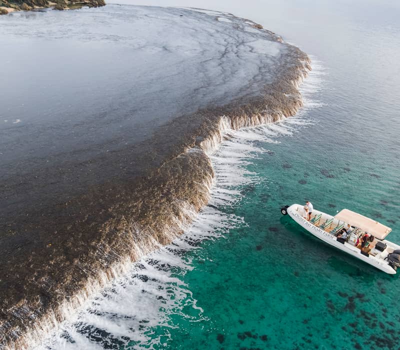 An aerial shot showing white water rushing over a dark coral reef shelf, creating a clear boundary with the turquoise ocean, with two tour boats nearby.
